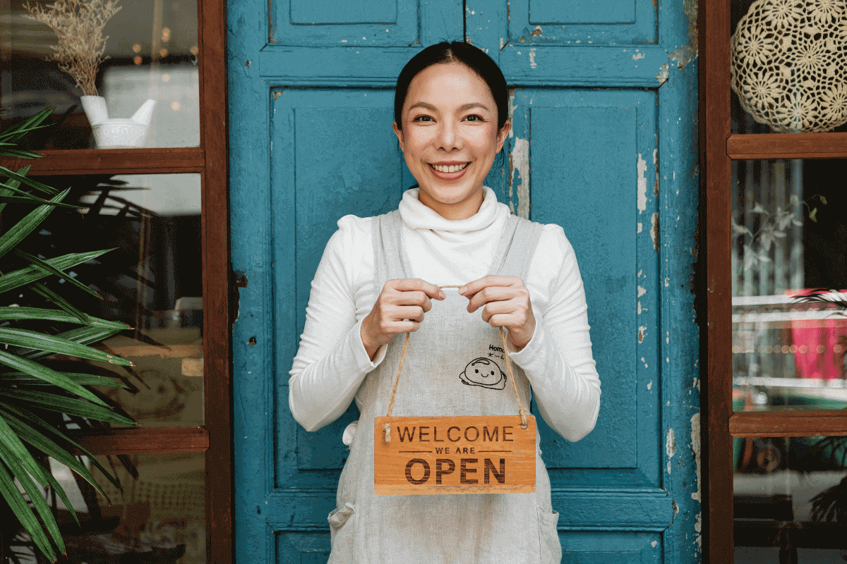 an asian female business owner standing outside her business with a welcome sign